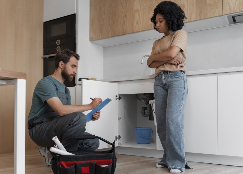 Plumber inspecting kitchen sink while homeowner watches during pre-sale home repair assessment in California