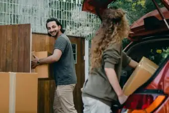 Cheerful modern male and female in comfortable casual clothes unpacking car trunk in green garden of new house while moving in together