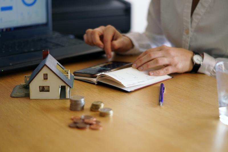 Person calculating housing costs with coins, notebook, and mini house.
