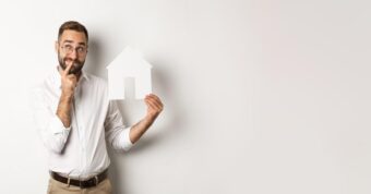 can-you-sell-your-house-after-a-year Man wondering while holding a paper house, “Can you sell your house after a year?”