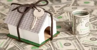 Mini house with key on dollar bills symbolizing home selling costs.