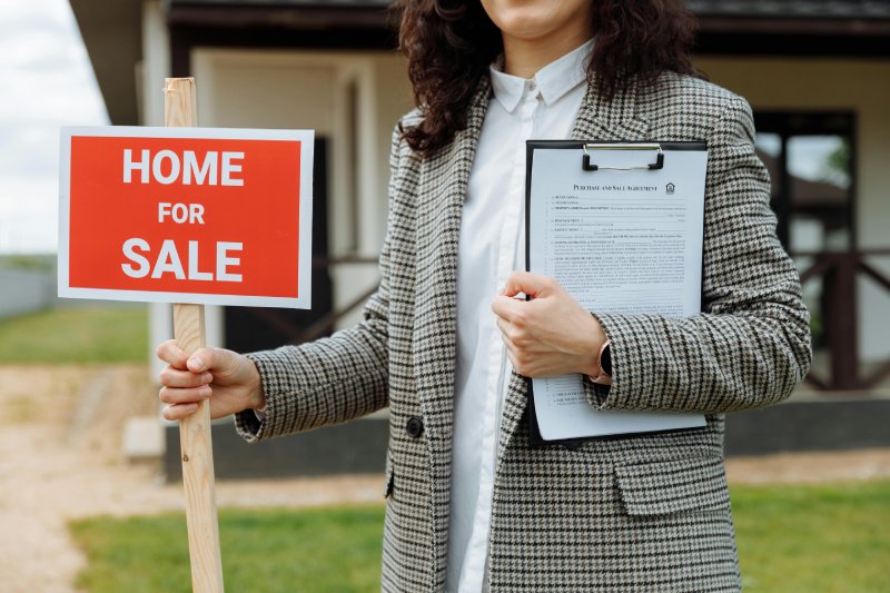 A woman holding a for sale sign in the front yard of a house.