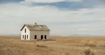 Abandoned white house with damaged roof in dry open field.