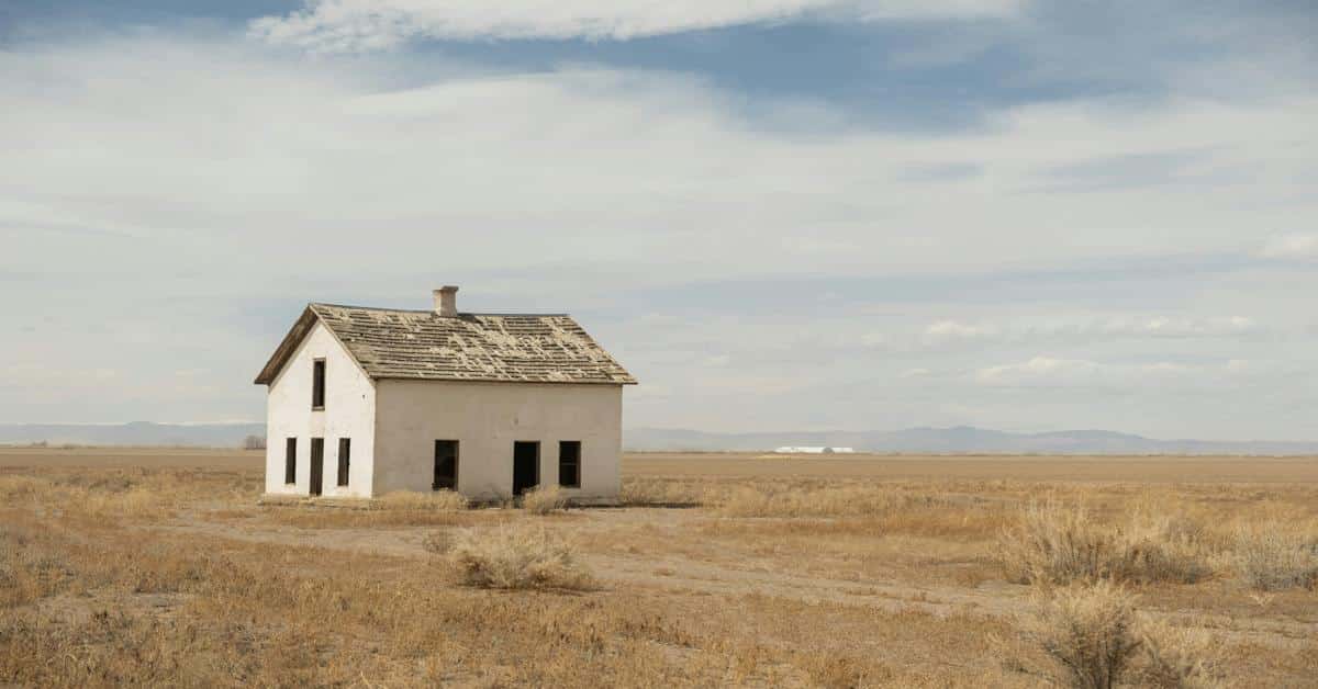 Abandoned white house with damaged roof in dry open field.
