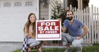 Smiling couple kneeling with For Sale By Owner sign