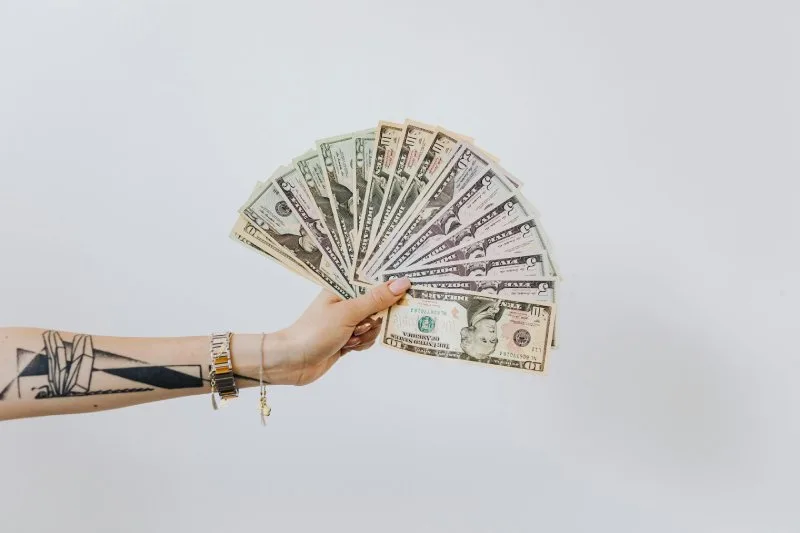 Tattooed arm holding mixed bills, showing lower-value cash offers