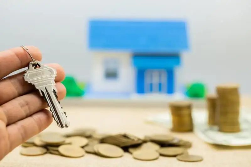Hand holding house key with money and coins in background.