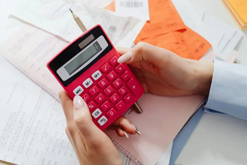 Hands holding red calculator over receipts and a financial paperwork stack.