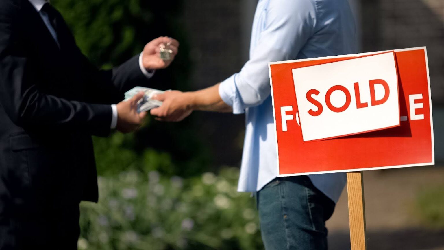 An estate agent and a man exchanging money and house keys against a sold signboard