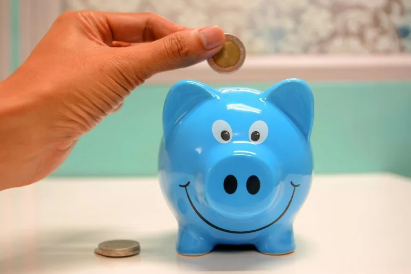 Hand placing coin into blue piggy bank to reduce costs.