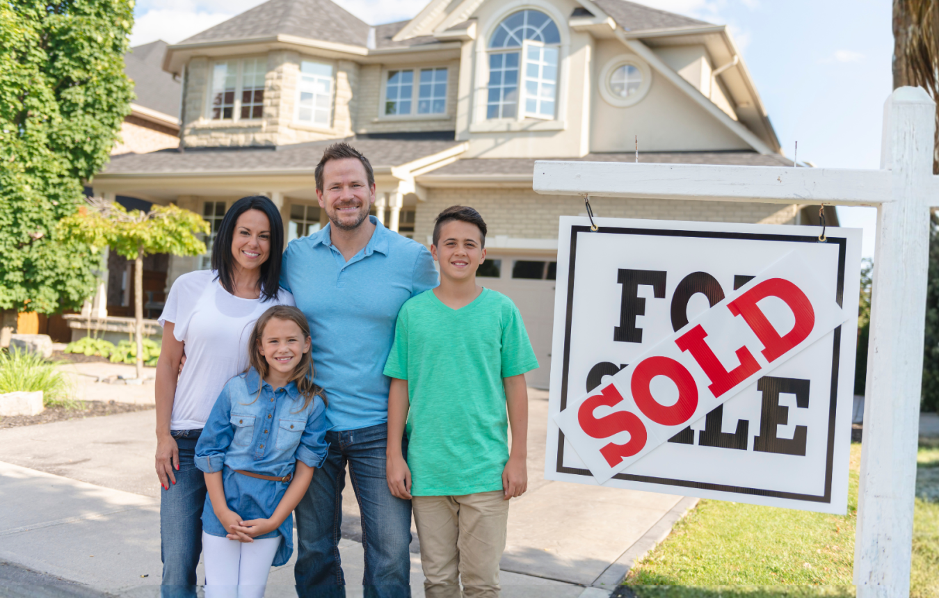 A family poses in front of their house with a "Sold" sign in the foreground.
